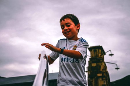 Boy In White And Blue Fly Emirates Jersey Shirt Holding On Stairs Grab Bar Under Gray Skies