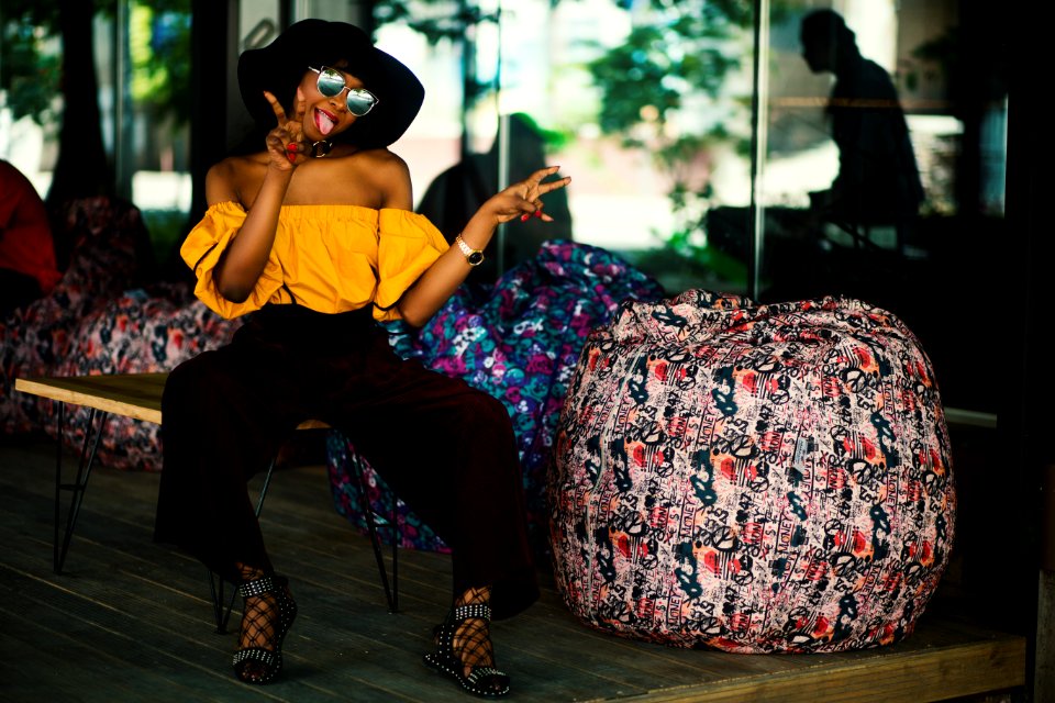 Woman Yellow Off-shoulder Top And Black Pants Sitting On Brown Bench With Tongue Out