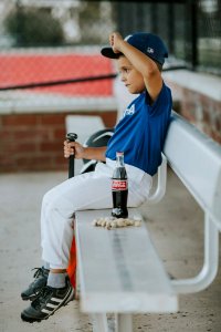 Selective Focus Photo Of A Boy Wearing Baseball Gear Sitting On Bench Beside Coca-cola Bottle