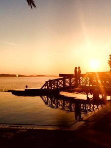 Silhouette Of Couple Standing Near Sea Dock At Golden Hour