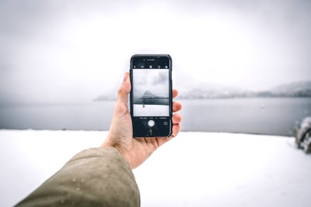 Photo Of Person Wearing Brown Coat Holding Android Smartphone While Taking Picture Of Mountain And Body Of Water photo