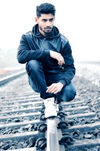 Man In Black Leather Zip-up Hoodie And Black Denim Jeans Sitting On Grey Metal Train Railings Surrounded With Rocks During Husky M photo