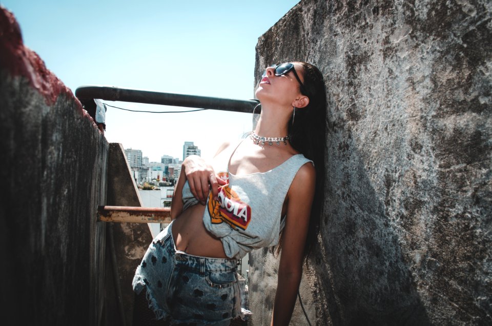 Woman Wearing White And Orange Tank Top And White Denim Shorts Leaning Of Gray Concrete Pavement