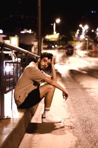 Man Wears Brown Shirt And Black Shorts Seats Beside Black Street Post