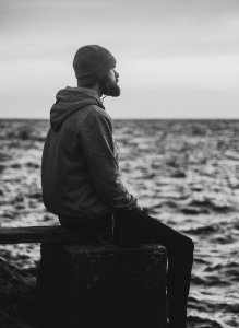 Grayscale Photo Of Man In Hoodie And Kit Cap Sitting Near Bodies Of Water photo