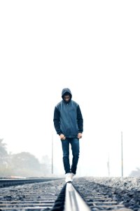 Man Wearing Gray And Black Zip-up Hoodie With Black Denim Jeans And White Shoes Walking On Train Railing Behind White Fog