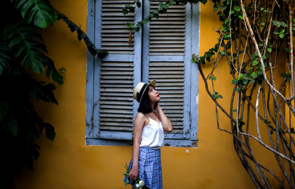 Photo Of Woman Wearing White Sleeveless Top And Hat Holding Flowers Near Window