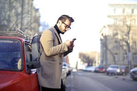 Man In Beige Coat Holding Phone Leaning On Red Vehicle