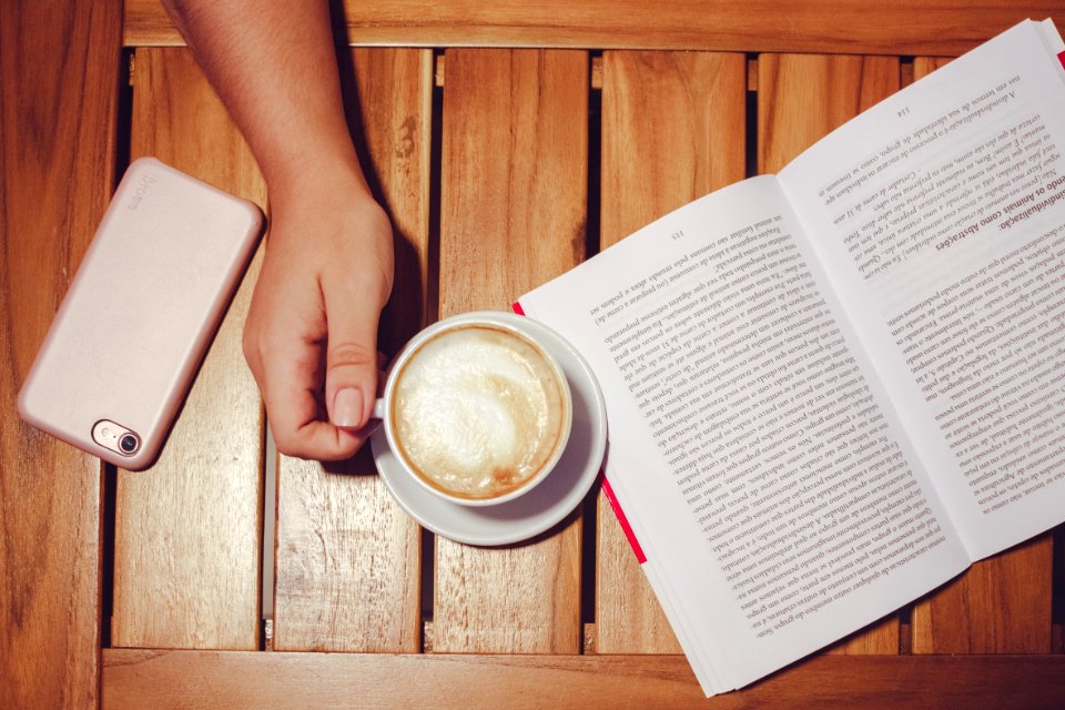 Persons Hand Holding White Coffee Mug With Plate On Brown Wooden Board With White And Black Book