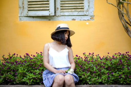 Woman In White Spaghetti Strapped Top With Blue And Purple Plaid Skirt Sitting On Ledge Near Purple Petaled Flower Garden Posing F