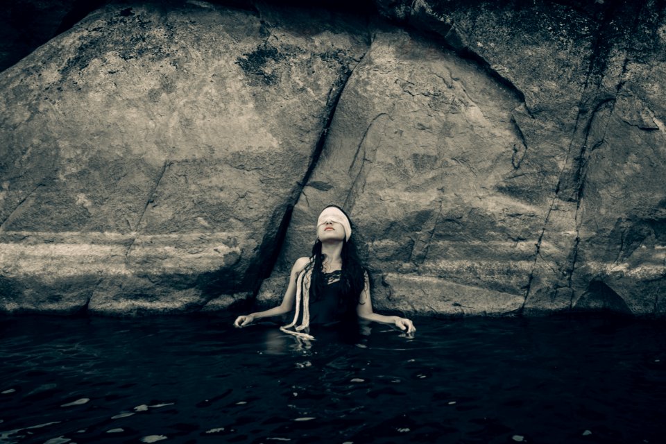 Woman In Blindfold Wearing Black Top On Body Of Water While Leaning On A Rock photo