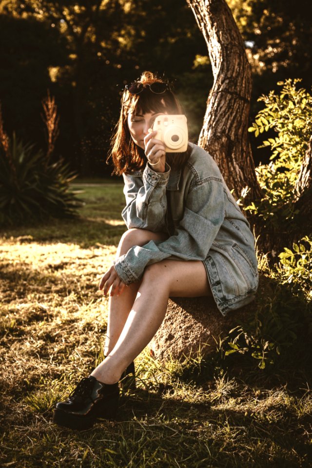 Woman Wearing Blue Denim Jacket Holding Pink Instant Camera Sitting On Soil photo