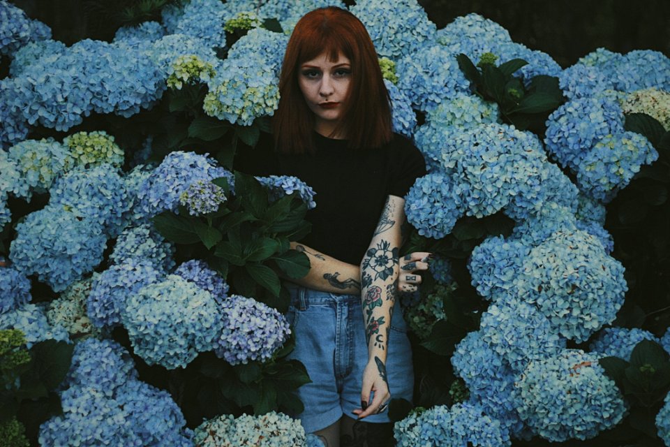 Woman With Black And Red Flower Tattoo Standing Behind Blue Flowers