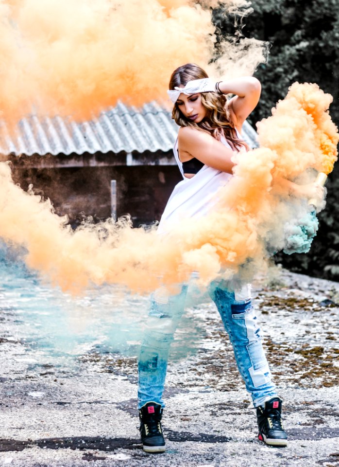 Woman Wearing White Tank Top And Blue Jeans Surrounded By Orange And Blue Smokes