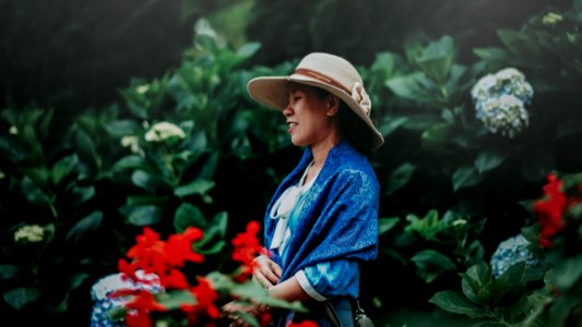 Selective Focus Photo Of Woman In Blue Shawl And Brown Sun Hat In The Middle Of Garden