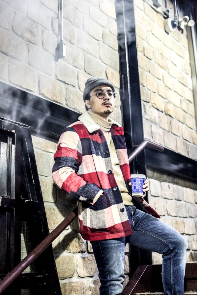 Man In Red Black And Grey Plaid Button-up Jacket With Blue Denim Jeans Outfit Holding Disposable Cup Stands Beside Brown Concret