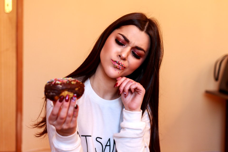 Woman In White Crew-neck Sweatshirt Holding And Looking At Chocolate Doughnut Inside Room