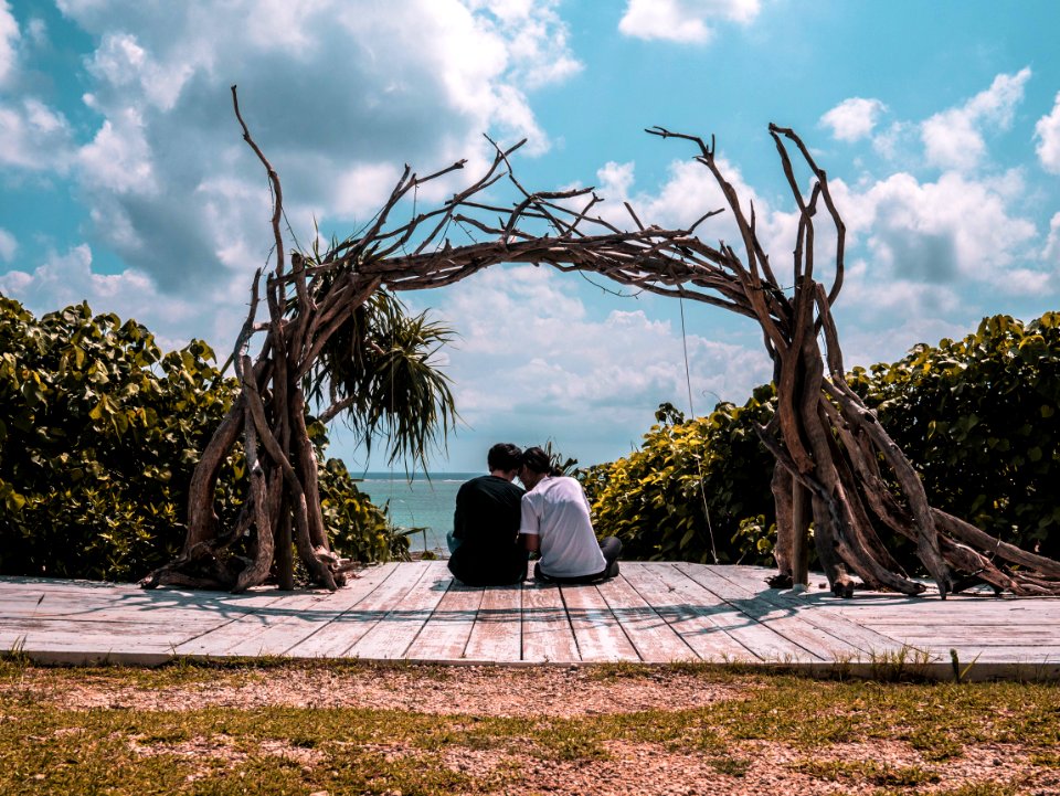Man And Woman Sitting On Wooden Dock Under Cloudy Sky