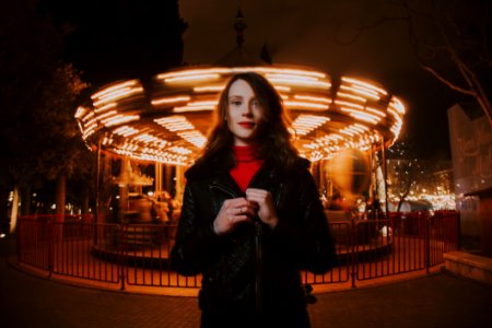 Woman Wearing Black Leather Jacket And Red Turtle-neck Shirt Standing In Front Of Carousel Ride