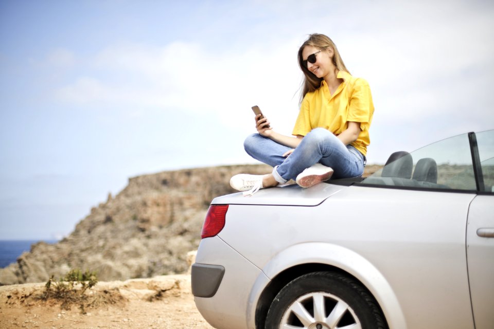 Woman In Yellow Blouse And Blue Jeans Taking Selfie While Sitting On Car