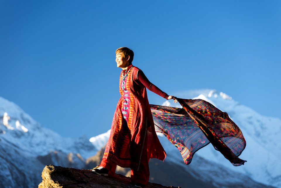 Woman In Orange Dress Standing On Top Of Rock Cliff Holding Scarf Near Mountain Covered With Snow
