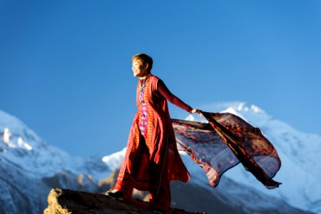 Woman In Orange Dress Standing On Top Of Rock Cliff Holding Scarf Near Mountain Covered With Snow photo