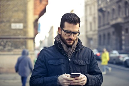 Man Wearing Black Zip Jacket Holding Smartphone Surrounded By Grey Concrete Buildings photo