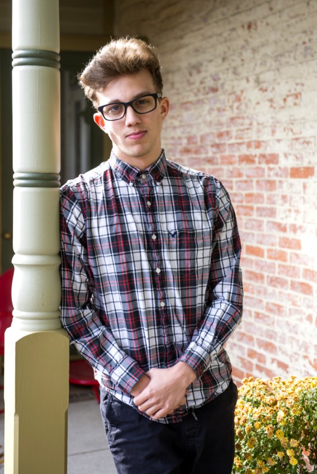 Blonde Man Wearing White Gray And Red Plaid Sport Shirt And Black Bottoms Standing While Leaning On Post