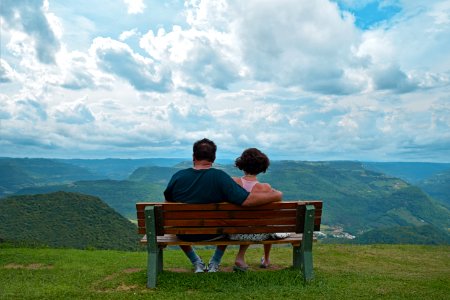 Couple Sitting On Brown Wooden Bench Near Mountains Covered With Grasses Under Blue Cloudy Sky