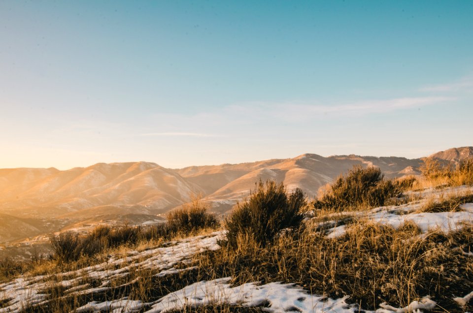 Green And Brown Grass Covered With Snow Overlooking Brown Hills And Mountains Under Clear Blue Sky At Daytime