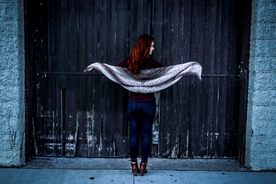 Woman Holding Gray Shawl While Spreading Her Arms Infront Of Brown Wooden Door