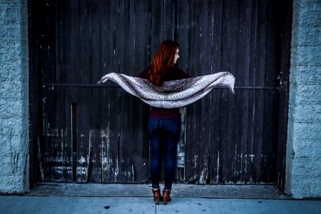 Woman Holding Gray Shawl While Spreading Her Arms Infront Of Brown Wooden Door
