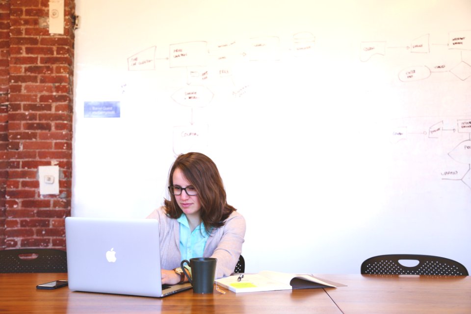 Woman Wearing Black Framed Eyeglasses And Teal Button-up Shirt And Beige Blazer Sitting At Table Near White Macbook