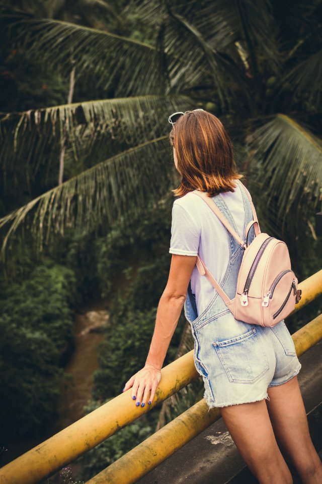 Woman Wearing Dungaree Shorts Stands Near A Yellow Metal Rail Overlook A River Belo With Coconut Trees At Daytime