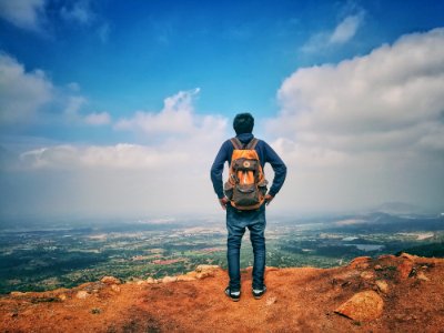 Man In Blue Dress Shirt And Blue Jeans And Orange Backpack Standing On Mountain Cliff Looking At Town Under Blue Sky And White Clo