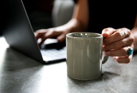 Person In Black Top Holding White Ceramic Mug And Using Laptop Computer photo