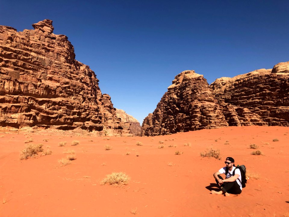 Man Wearing White Shirt And Black Pants Sitting On Soil Behind Rock Formation Mountain