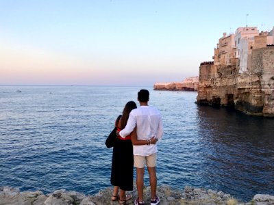 Man In White Dress Shirt Standing Beside The Woman In Black And Red Dress While Watching The Blue Calm Water Near Brown Concrete B photo