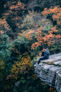 Photography Of Man Wearing Black Hoodie With Black Pants Sitting On Stone Cliff Above Green And Red Leaved Forest photo
