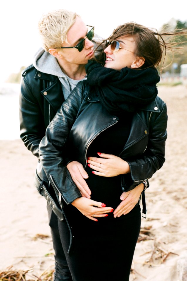 Man In Black Leather Zip Jacket Hugging Woman In Black Leather Zip Jacket And Black Pants Standing On Sand At Daytime