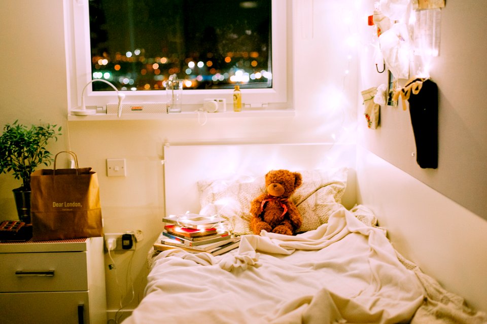 Brown Bear Plush Toy On White Bed Comforter Inside Lighted Bedroom