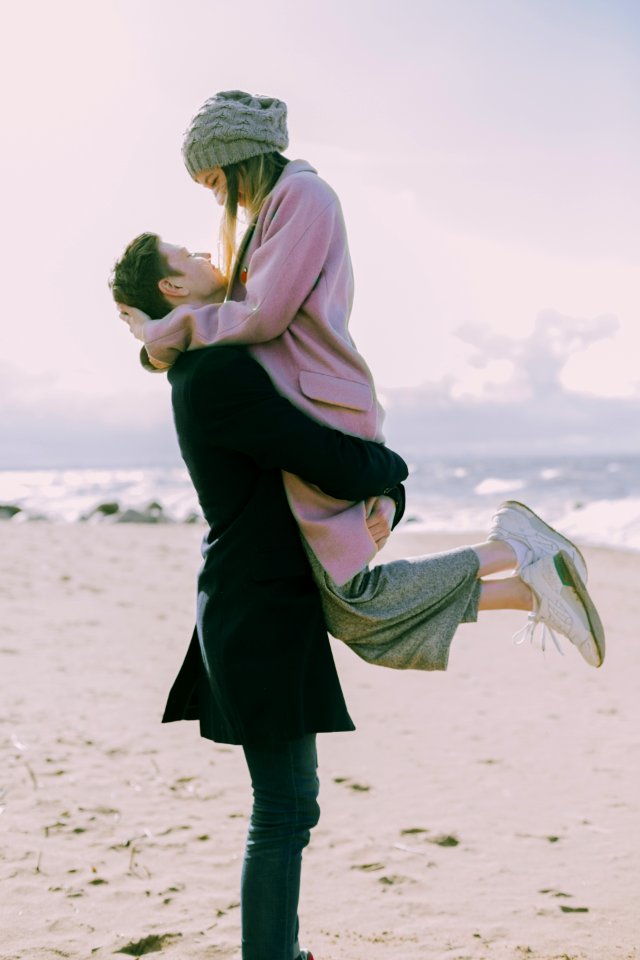 Man In Gray Coat Carrying Woman Wearing Pink Coat In Beach Near Shoreline And Body Of Water