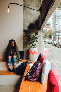 Woman In Gray Zip Up Jacket And Blue Distressed Jeans Sitting On Bench Near Backpack Looking Outside In Room photo