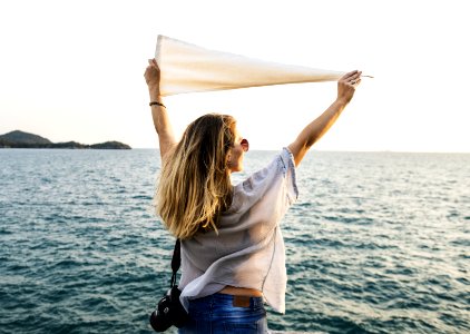 Photo Of Woman Wearing White Top Blue Bottoms And Black Dslr Camera Holding White Textile While Facing The Ocean photo