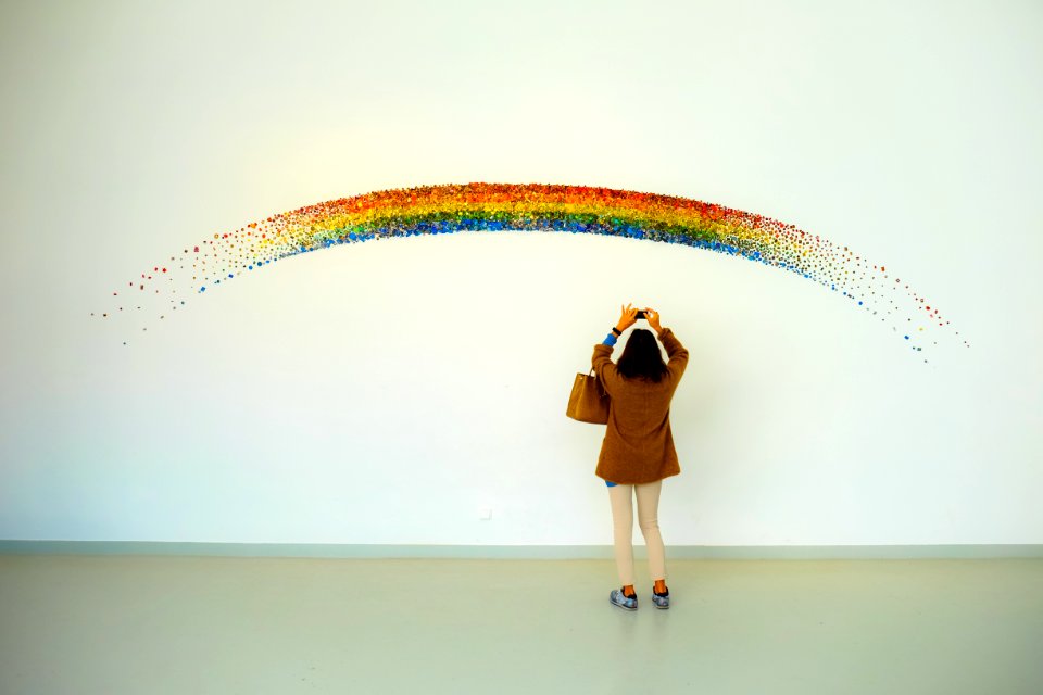 Woman Wearing Brown Top And Beige Leggings Taking Picture Rainbow Painted Wall