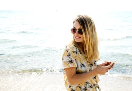 Woman In Black Yellow And White Floral Button-up Shirt Holding Smartphone Wearing Aviator Sunglasses Near Body Of Water photo