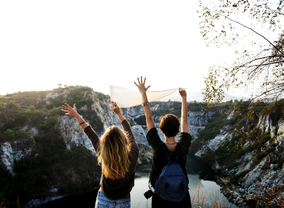 Man Holding White Pennant Beside Girl Raising Two Hands In Front Of Lake Surrounded By Mountains