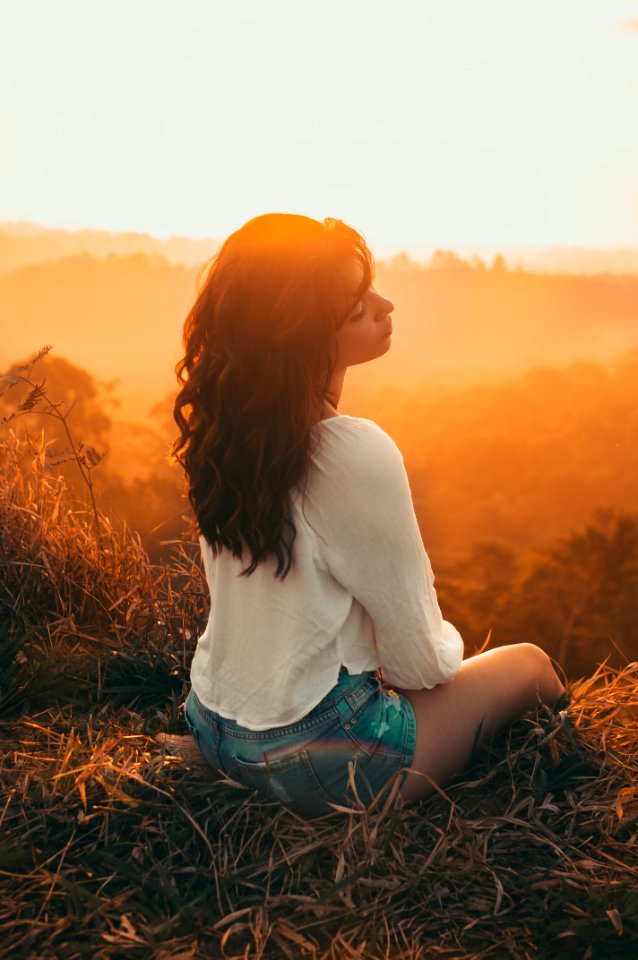 Woman In White Long-sleeved Shirt With Blue Short Shorts Sitting On Brown Grass Against Golden Hour Light