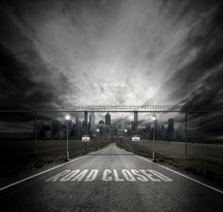 Grayscale Photo Of Road Closed On Roadway With Gray Fence Gate In Front Of City