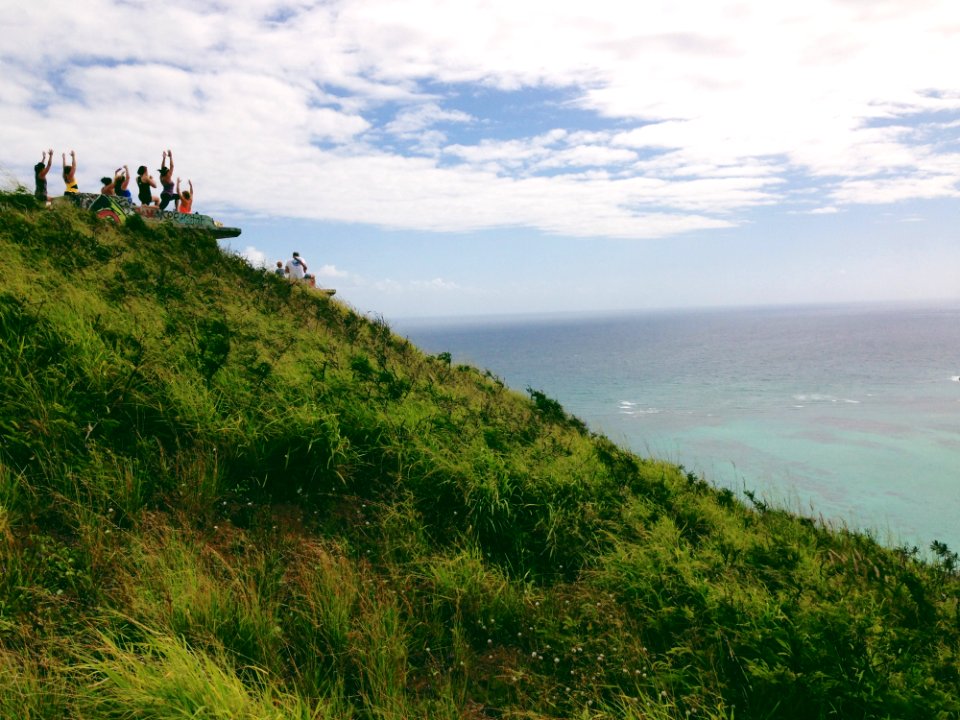 People Standing On A Green Mountain Peak Watching The Clear Blue Body Of Water